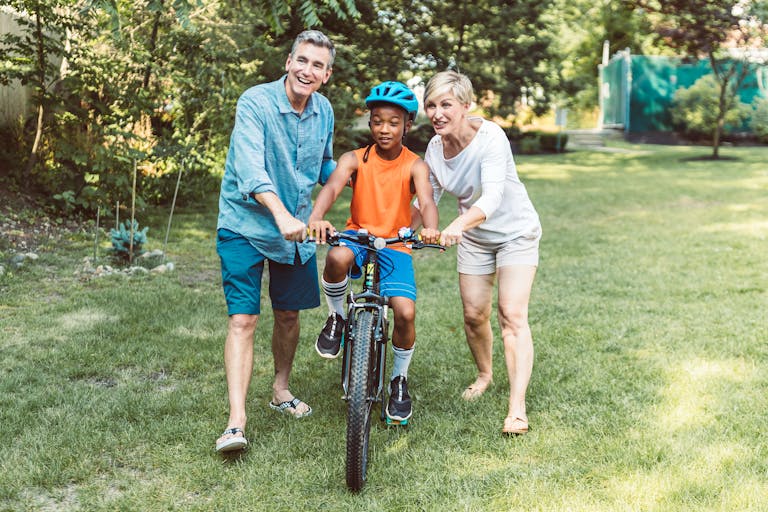 A family helps a young boy learn to ride a bicycle outdoors.